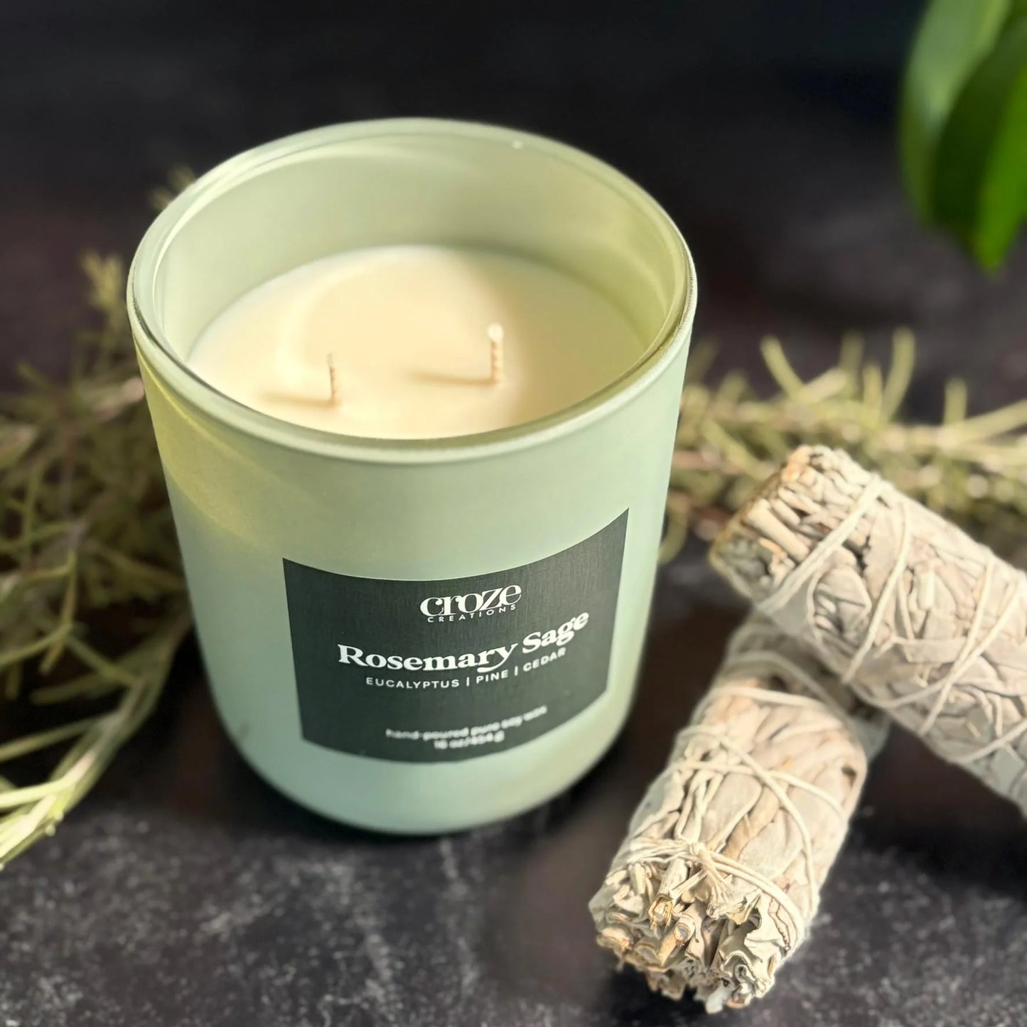 Candle labeled 'Rosemary Sage' with a smudge stick on a dark surface