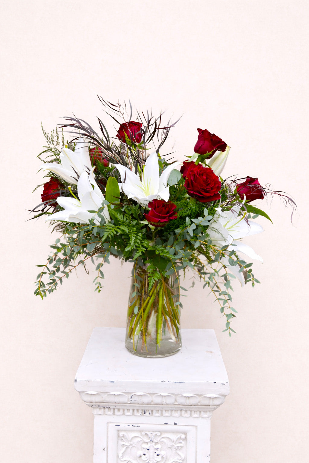 Bouquet of red roses and white lilies in a clear vase on a white pedestal against a light background